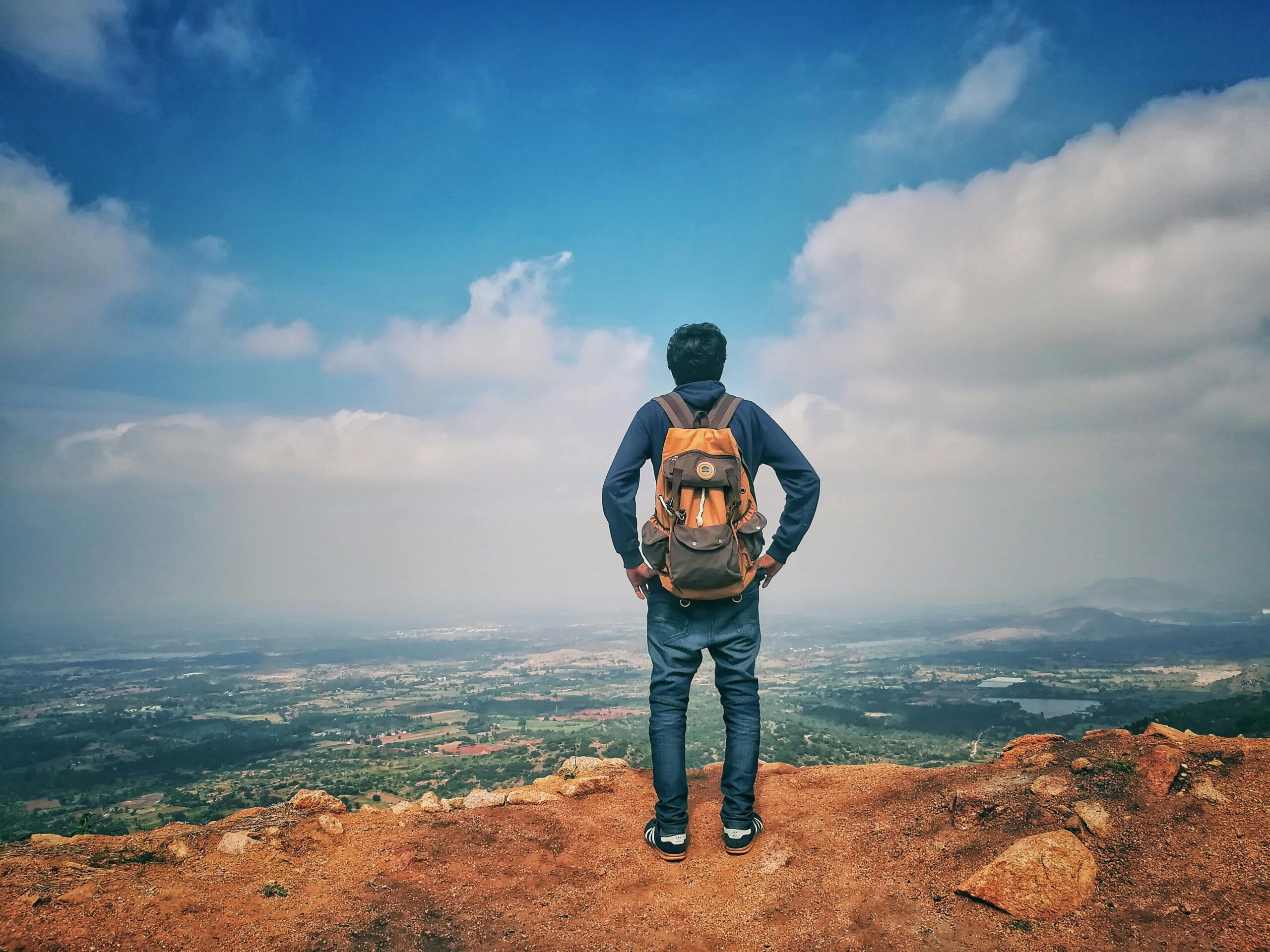 A man stands on a mountain edge appreciating the expansive scenic view below under a bright sky.